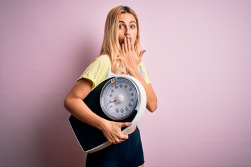 Young beautiful blonde sporty woman on diet holding weight machine over pink background cover mouth with hand shocked with shame for mistake, expression of fear, scared in silence, secret concept