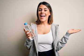 Young beautiful brunette sporty woman drinking bottle of water over isolated white background very happy and excited, winner expression celebrating victory screaming with big smile and raised hands