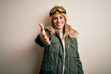 Young brunette skier woman wearing snow clothes and ski goggles over white background smiling friendly offering handshake as greeting and welcoming. Successful business.