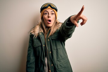 Young brunette skier woman wearing snow clothes and ski goggles over white background Pointing with finger surprised ahead, open mouth amazed expression, something on the front
