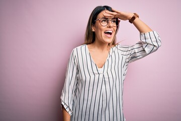 Young beautiful woman wearing casual striped t-shirt and glasses over pink background very happy and smiling looking far away with hand over head. Searching concept.