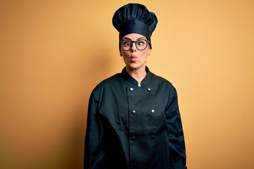 Young beautiful brunette chef woman wearing cooker uniform and hat over yellow background making fish face with lips, crazy and comical gesture. Funny expression.