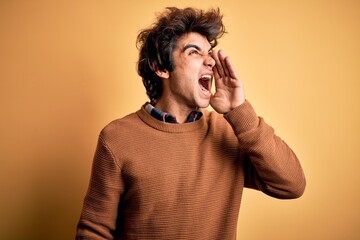 Young handsome man wearing casual shirt and sweater over isolated yellow background shouting and screaming loud to side with hand on mouth. Communication concept.