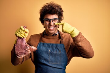 Young handsome butcher man holding meet steak standing over isolated yellow background Pointing with hand finger to face and nose, smiling cheerful. Beauty concept