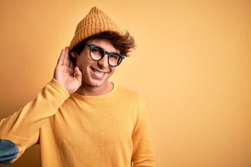 Young handsome man wearing casual t-shirt and glasses over isolated yellow background smiling with hand over ear listening an hearing to rumor or gossip. Deafness concept.