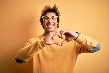Young handsome man wearing casual t-shirt and glasses over isolated yellow background smiling in love showing heart symbol and shape with hands. Romantic concept.