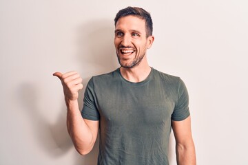 Young handsome man wearing casual t-shirt standing over isolated white background pointing thumb up to the side smiling happy with open mouth