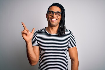 Young handsome african american man with dreadlocks wearing striped t-shirt and glasses with a big smile on face, pointing with hand and finger to the side looking at the camera.