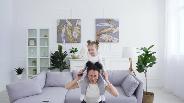 Front view of appealing fit smiling active young woman which doing squat exercises with her funny cute small daughter on her shoulders in the living-room