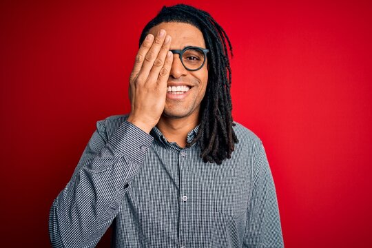 Young handsome african american man with dreadlocks wearing casual shirt and glasses covering one eye with hand, confident smile on face and surprise emotion.