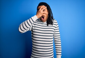 Young handsome african american afro man with dreadlocks wearing casual striped sweater peeking in shock covering face and eyes with hand, looking through fingers with embarrassed expression.