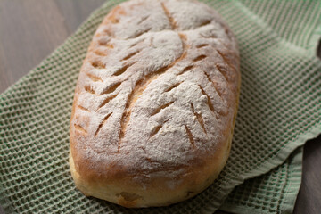 Homemade artisan sourdough bread loaf with artistic leaf pattern scoring and a wavy line in the middle on a green kitchen towel on wooden surface. Rustic aesthetic 