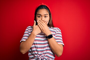 Young beautiful asian girl wearing casual striped t-shirt over isolated red background shocked covering mouth with hands for mistake. Secret concept.