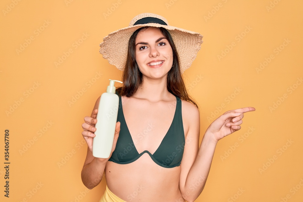 Poster Young beautiful girl wearing swimwear bikini and summer hat holding sun protection cream very happy pointing with hand and finger to the side