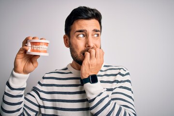 Young handsome man with beard holding plastic denture teeth over white background looking stressed and nervous with hands on mouth biting nails. Anxiety problem.