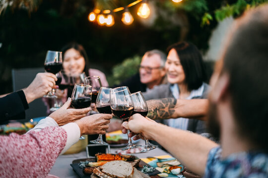 Happy family cheering with red wine at barbecue dinner outdoor at home - Different age of people having fun at weekend meal - Food, taste and summer concept - Focus on on left bottom hand