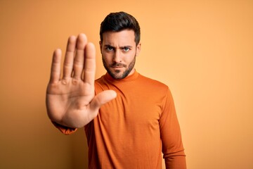 Young handsome man with beard wearing casual sweater standing over yellow background doing stop sing with palm of the hand. Warning expression with negative and serious gesture on the face.