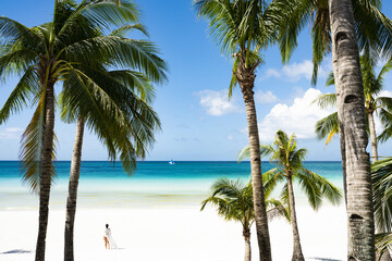 Fototapeta premium (Selective focus) Stunning view of woman walking on a white sand beach bathed by a turquoise sea, beautiful coconut palm trees in the foreground. White Beach, Boracay Island, Philippines.