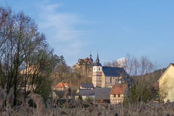 Dreifaltigkeitskirche mit Turm aus Schiefer am Schlossberg in Neuhaus-Schierschnitz in Thüringen. 
Kirche am Berg neben einer Burg bei blauem Himmel mit Bäumen und Steinhäusern.