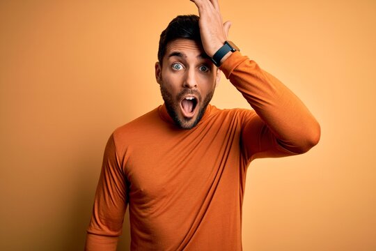Young handsome man with beard wearing casual sweater standing over yellow background surprised with hand on head for mistake, remember error. Forgot, bad memory concept.