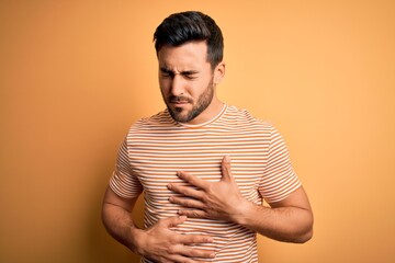 Young handsome man with beard wearing casual striped t-shirt over yellow background with hand on stomach because indigestion, painful illness feeling unwell. Ache concept.