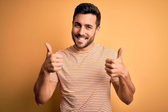 Young handsome man with beard wearing casual striped t-shirt over yellow background success sign doing positive gesture with hand, thumbs up smiling and happy. Cheerful expression and winner gesture.