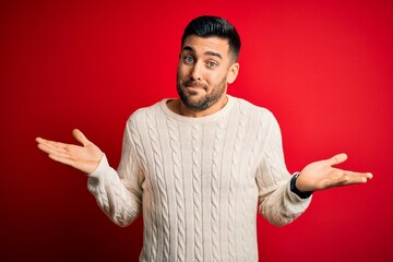 Young handsome man wearing casual white sweater standing over isolated red background clueless and confused expression with arms and hands raised. Doubt concept.