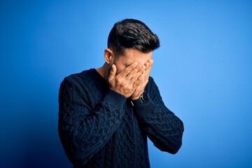 Young handsome man wearing casual sweater standing over isolated blue background with sad expression covering face with hands while crying. Depression concept.