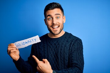 Young handsome man holding paper with solidarity message over blue background very happy pointing...
