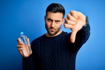 Young handsome man drinking bottle of water to refeshment over blue background with angry face,...