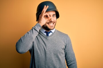 Young handsome businessman wearing military helmet over isolated yellow background doing ok gesture with hand smiling, eye looking through fingers with happy face.