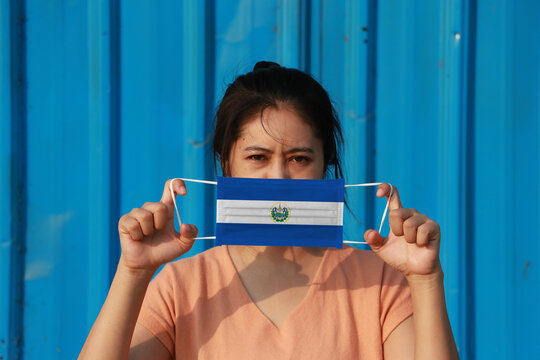 A Woman With El Salvador Flag On Hygienic Mask In Her Hand And Lifted Up The Front Face On Blue Background. Tiny Particle Or Virus Corona Or Covid 19 Protection.