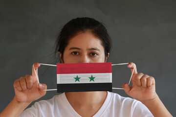 A woman in white shirt with Syria flag on hygienic mask in her hand and lifted up the front face on dark grey background. Tiny Particle or virus corona or Covid 19 protection.