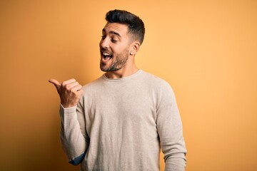 Young handsome man wearing casual sweater standing over isolated yellow background smiling with happy face looking and pointing to the side with thumb up.