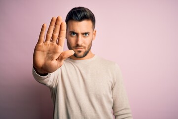 Young handsome man wearing casual sweater standing over isolated pink background doing stop sing with palm of the hand. Warning expression with negative and serious gesture on the face.