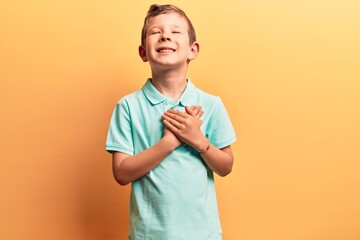 Cute blond kid wearing casual clothes smiling with hands on chest, eyes closed with grateful...