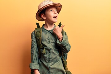 Cute blond kid wearing explorer hat and backpack pointing thumb up to the side smiling happy with open mouth