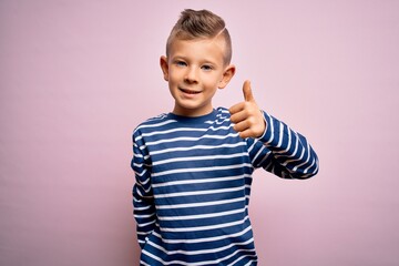 Young little caucasian kid with blue eyes wearing nautical striped shirt over pink background doing happy thumbs up gesture with hand. Approving expression looking at the camera showing success.