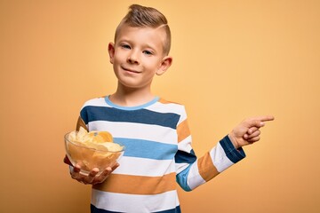 Young little caucasian kid eating unheatlhy potatoes crisps chips over yellow background very happy...