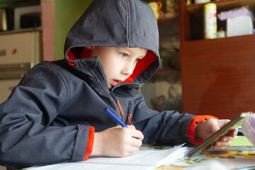 A European teen boy in a jacket sits at a table and does homework. Homework from school. It's cold...
