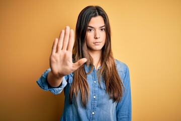 Young beautiful girl wearing casual denim shirt standing over isolated yellow background doing stop sing with palm of the hand. Warning expression with negative and serious gesture on the face.