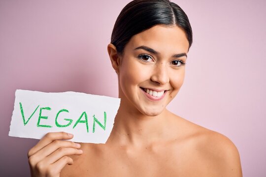 Young beautiful girl holding paper with vegan message over isolated pink background with a happy face standing and smiling with a confident smile showing teeth