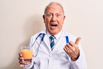 Senior grey-haired doctor man wearing stethoscope drinking glass of healthy orange juice pointing thumb up to the side smiling happy with open mouth
