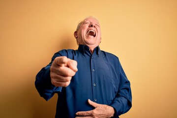 Grey haired senior man wearing casual blue shirt standing over yellow background laughing at you,...
