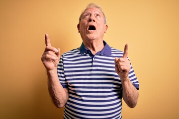 Grey haired senior man wearing casual navy striped t-shirt standing over yellow background amazed and surprised looking up and pointing with fingers and raised arms.