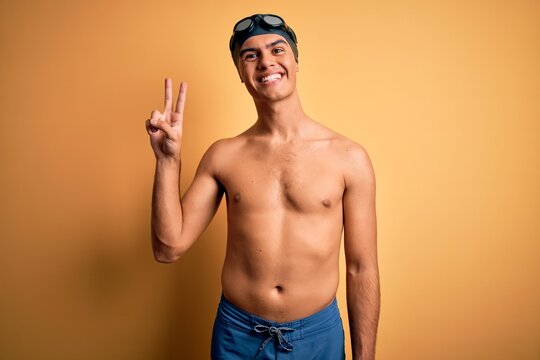 Young handsome man shirtless wearing swimsuit and swim cap over isolated yellow background showing and pointing up with fingers number two while smiling confident and happy.