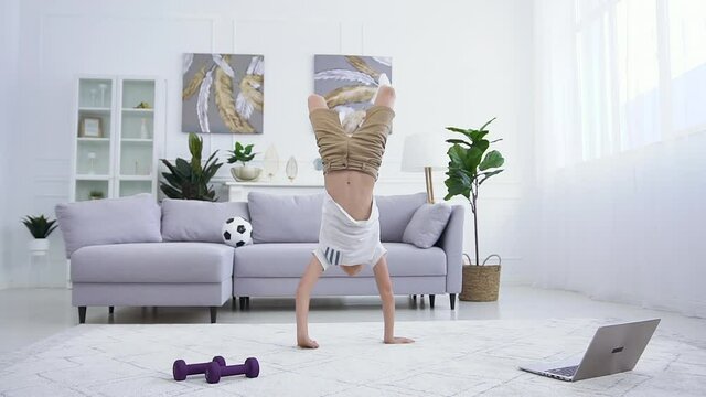 Good-looking sportive confident teen boy doing handstand on the carpet,walking on his hands and falling down on the couch
