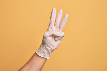 Hand of caucasian young man with medical glove over isolated yellow background counting number 3 showing three fingers