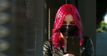 Caucasian young stylish hipster with pink hair tapping or texting message on smartphone outdoors. Extraordinary girl in mask scrolling on mobile phone. Female using cellphone. Covid-19 pandemic.