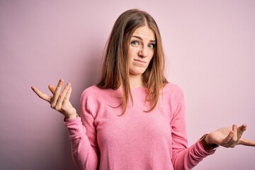 Young beautiful redhead woman wearing casual sweater over isolated pink background clueless and confused with open arms, no idea concept.